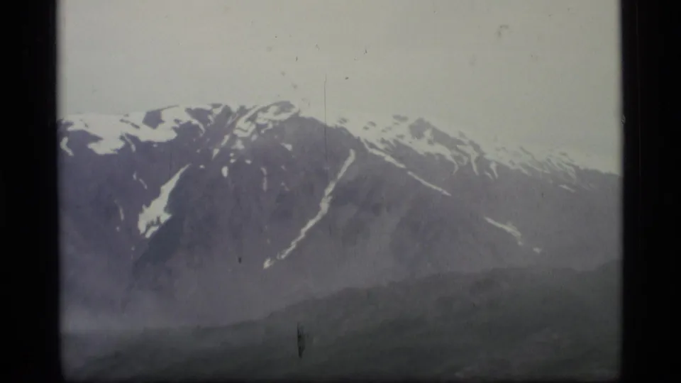 1977 Alaska 8mm frame showing a broad mountain face with irregular snow patches, muted haze, and dark valley foreground.