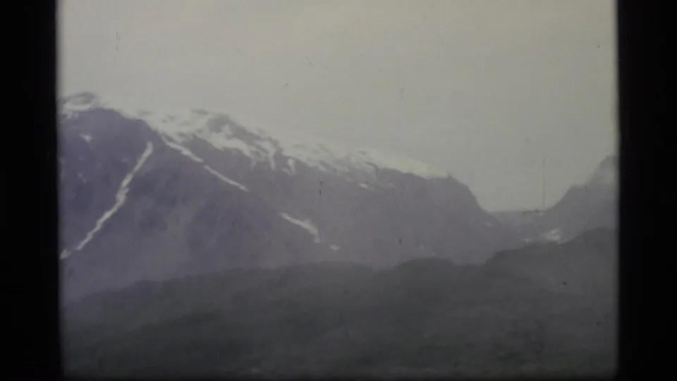 1977 Alaska 8mm frame of a mountain ridge receding into mist, with snow streaks on upper slopes and a pale sky.