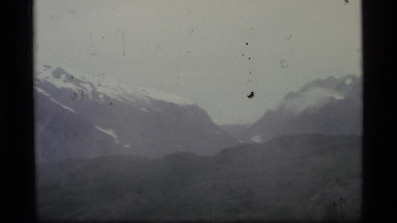 1977 Alaska 8mm frame showing distant snow-capped mountains, low cloud, and a shadowed valley in soft faded color.