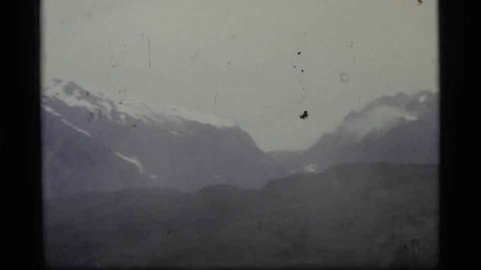 1977 Alaska 8mm frame showing distant snow-capped mountains, low cloud, and a shadowed valley in soft faded color.