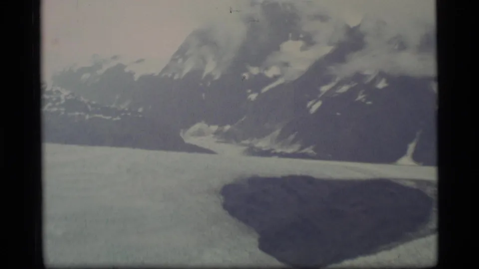 1977 Alaska 8mm frame showing pale glacier ice or an ice field beneath steep mountains and clouded snow patches.