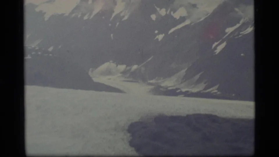 1977 Alaska 8mm view of a glacier edge beneath a steep snow-streaked mountain slope, with dark terrain in the foreground.