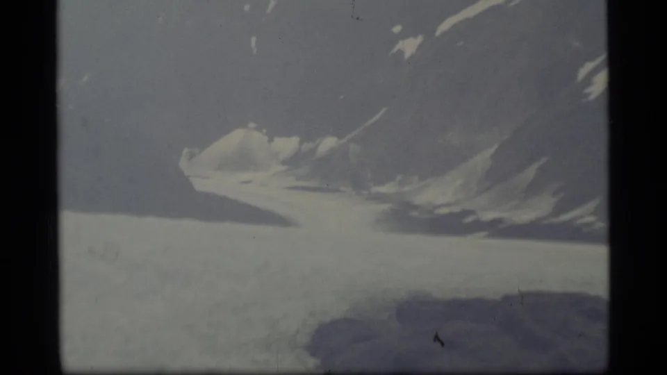 1977 Alaska 8mm frame showing a pale glacier surface and snow-topped mountain ridge beneath an overcast sky.