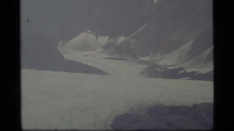 1977 Alaska home-movie frame with a broad glacier foreground and a snow-lined mountain ridge receding into haze.