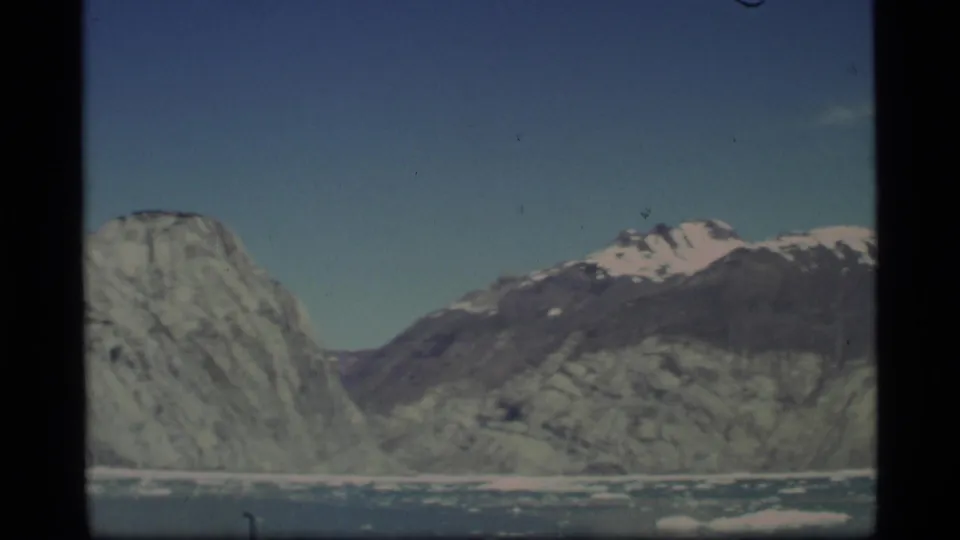 1977 Alaska 8mm frame with blue sky, snow-dusted peaks, steep valley walls, and glacier ice in the foreground.