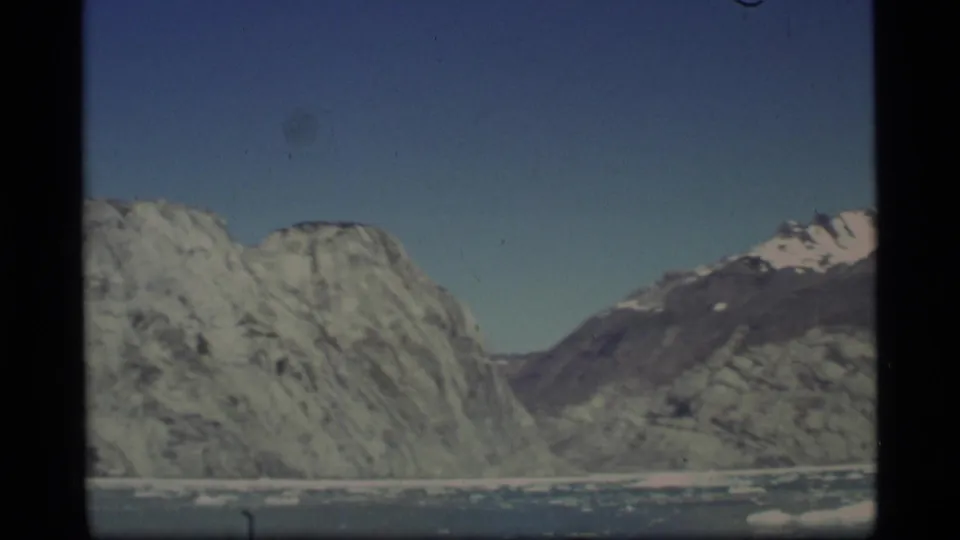 1977 Alaska 8mm frame showing rugged snow-dusted mountains across icy water with scattered floes and a clear sky.
