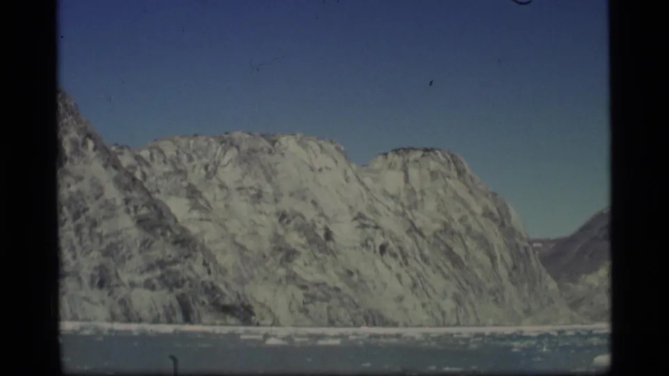 1977 Alaska 8mm frame with a broad rocky mountain wall, snow traces, blue sky, and ice floes on the water below.