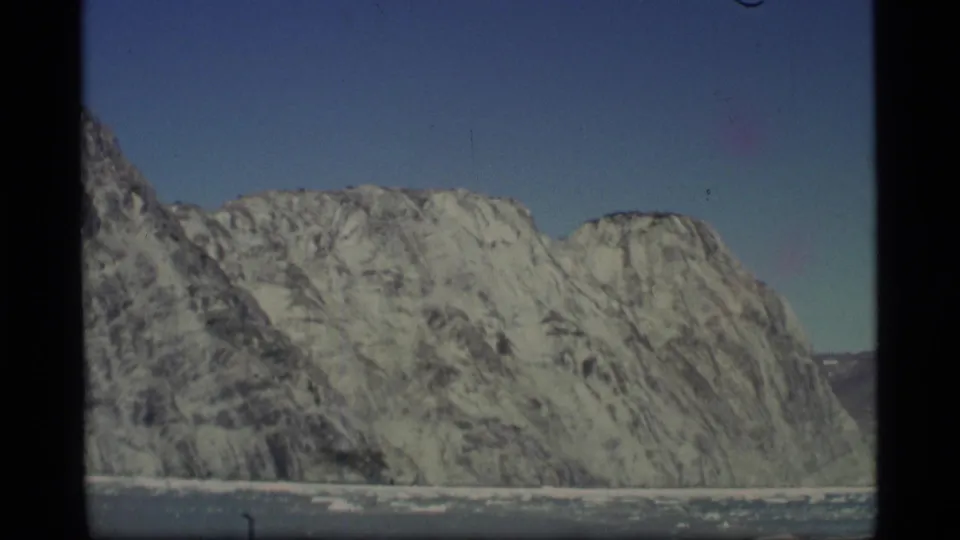 1977 Alaska 8mm frame showing steep snow-streaked mountain slopes meeting icy water with scattered floes in the foreground.