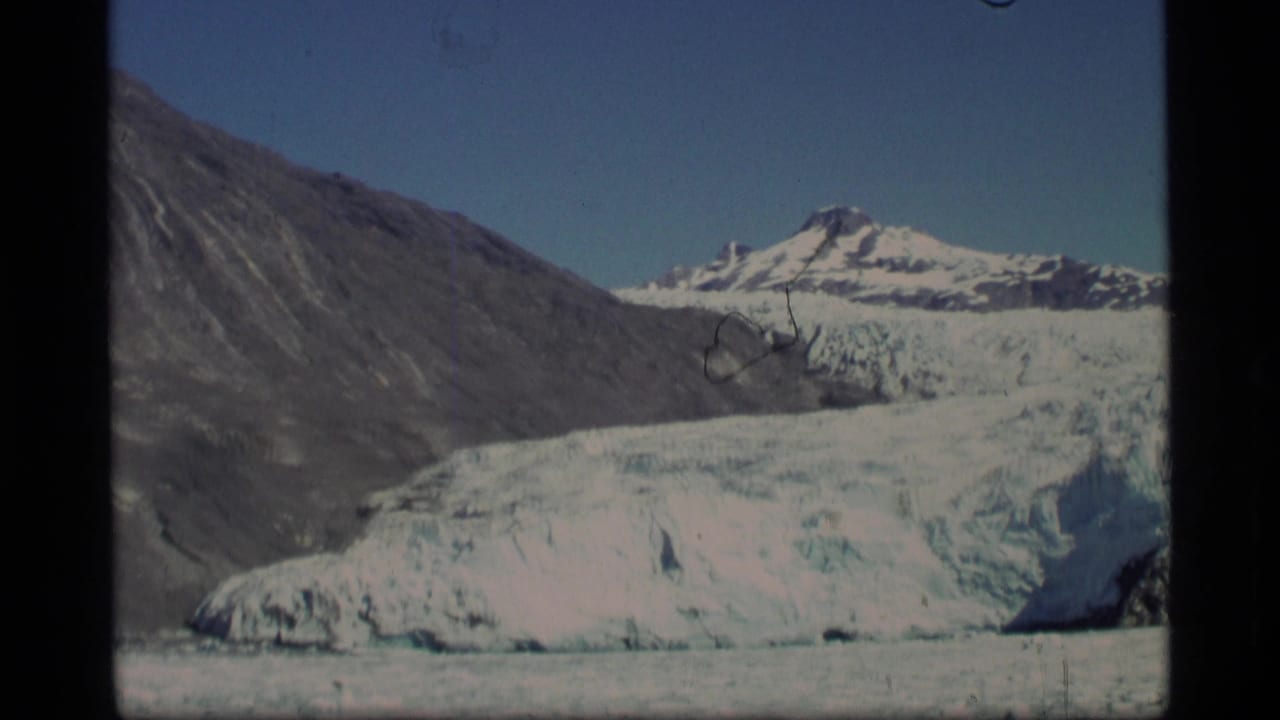 1977 Alaska 8mm frame showing a blue-white glacier below rocky mountain slopes under a clear pale sky.