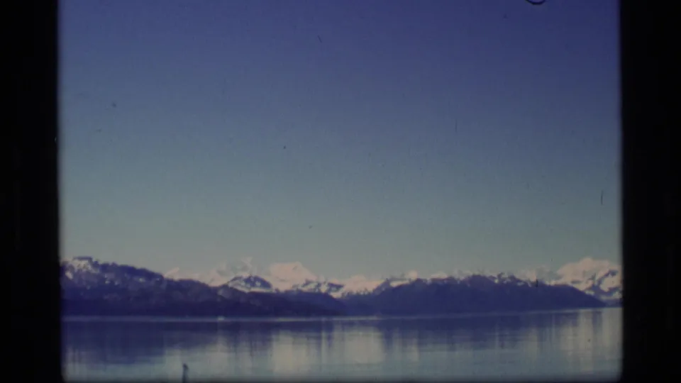 1977 8mm Alaska view across a still lake toward distant snow-capped mountains under blue sky.