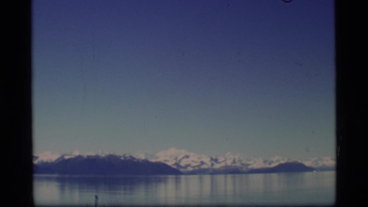 Snow-capped mountains across calm water under a blue sky, seen within dark film-frame borders.