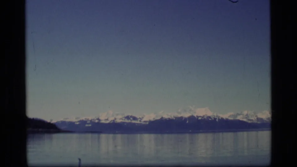 1977 home-movie view of an Alaskan mountain range across calm water, with a dark shoreline edge visible at left.