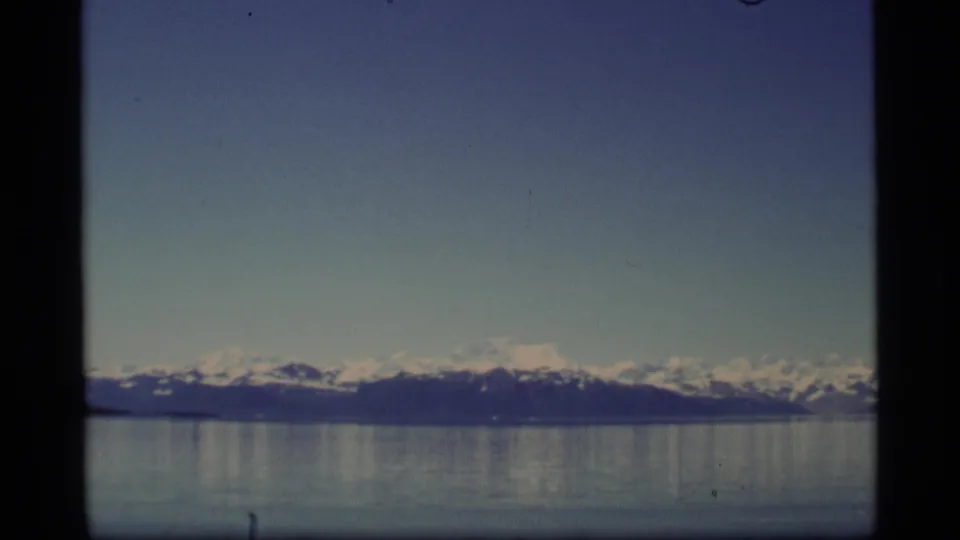 Alaska 8mm footage frame with distant snow-capped mountains and muted reflections on smooth water.