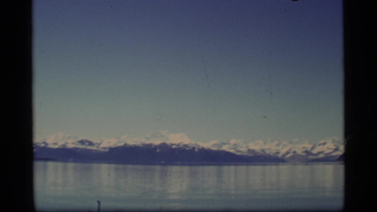 Snow-capped mountains across calm water under a blue sky.