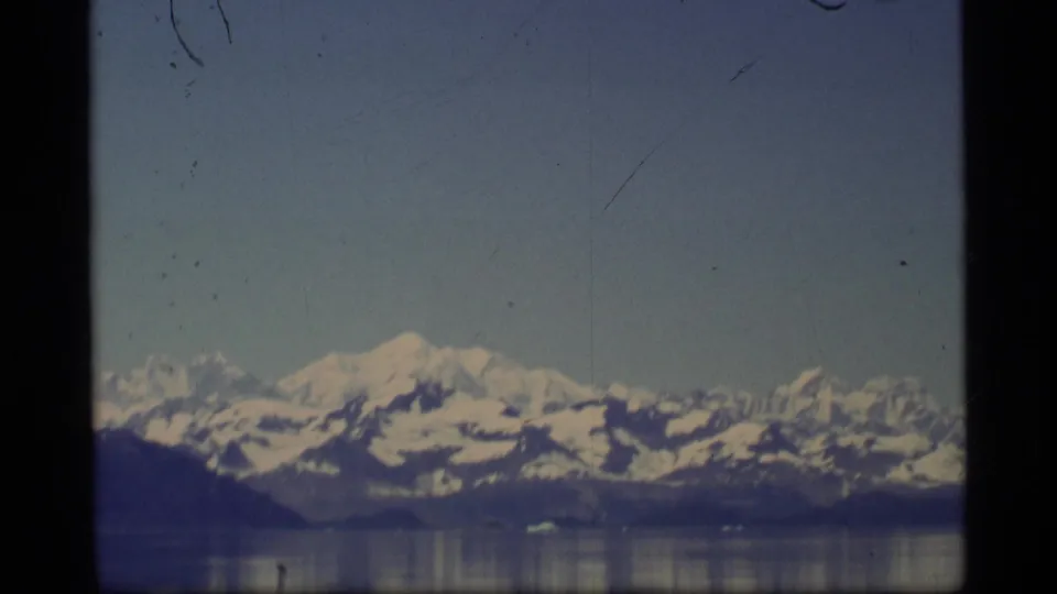 1977 Alaska home-movie view of a snow-covered mountain range above calm water, framed by visible film edges.