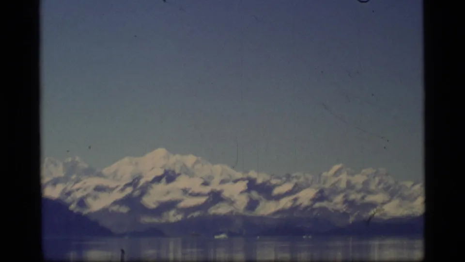 1977 Alaska Super 8 frame with snow-capped mountains, a dark shoreline, and calm reflective water.
