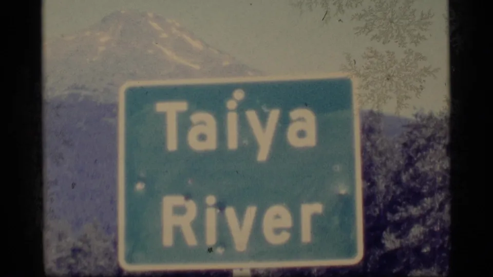 1977 Alaska frame showing a Taiya River road sign in front of a faint mountain backdrop and roadside vegetation.