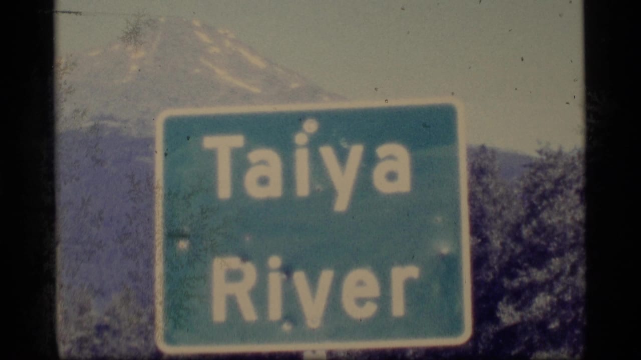 1977 Alaska 8mm frame showing a green Taiya River sign in front of trees and distant mountain slopes.