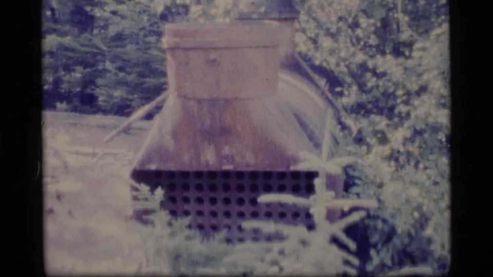 1977 Alaska Super 8 frame centered on a rusting metal structure with a perforated lower section and surrounding forest plants.