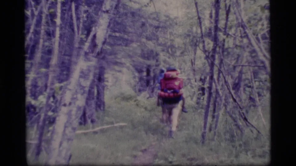A hiker with a red backpack walks away through an Alaska forest trail in faded 1970s 8mm footage.