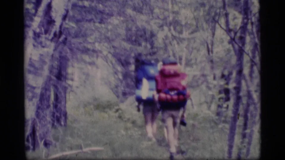 Two backpacked hikers walk away along a wooded Alaska trail, filmed from behind in 1977 8mm home-movie style.