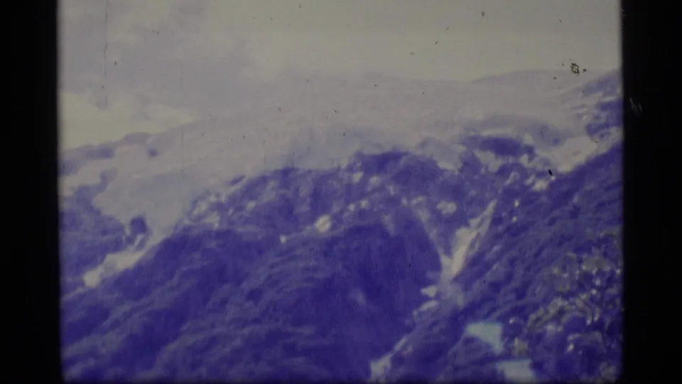 1977 Alaska 8mm frame showing a distant mountain ridge with snow patches, pale clouds, and strong blue-purple film color shift.