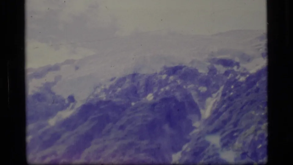 1977 Alaska 8mm frame with soft, blue-toned mountain slopes, lingering snow, pale sky, and visible film-frame edge.