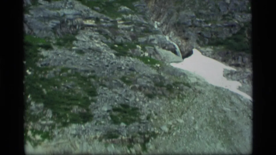 1977 Alaska 8mm frame showing rocky alpine terrain with green shrub growth, a white melting snow patch, and handheld home-movie texture.