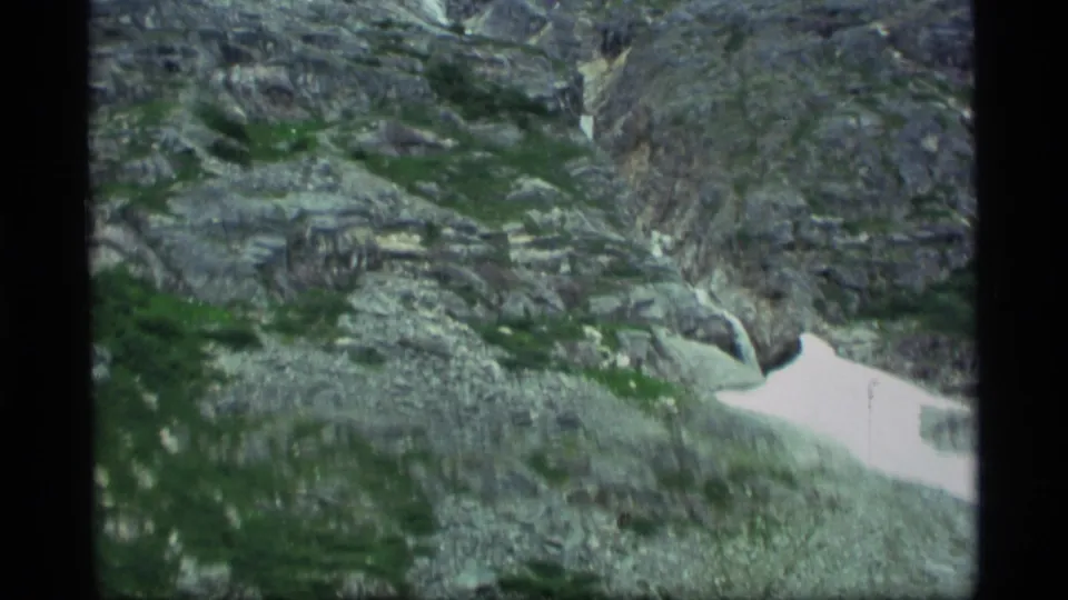 1977 Alaska 8mm frame of rugged gray rock, scattered green vegetation, and a bright remaining snow patch on the lower slope.