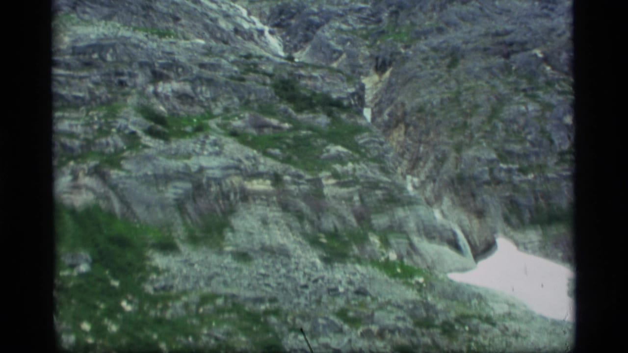 1977 Alaska 8mm aerial view of gray mountain rock, green vegetation, and a bright snow patch near a ravine.