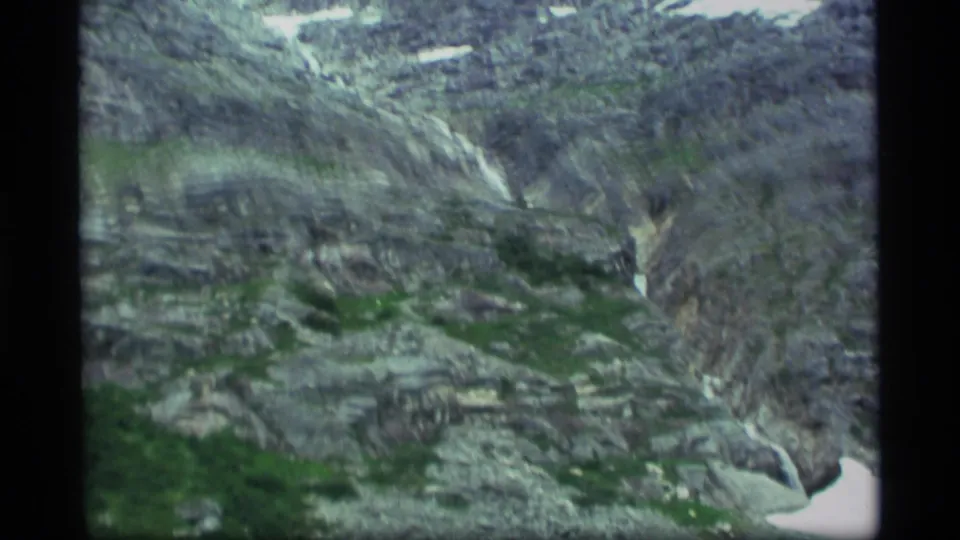 1977 Alaska aerial home-movie frame showing striated rocky slopes, scattered green vegetation, and snow on high ground.