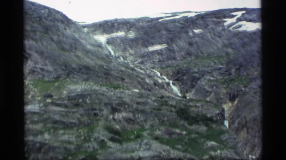 1977 Alaska 8mm aerial frame of a rocky mountain valley with a thin stream or waterfall below snow-streaked ridges.