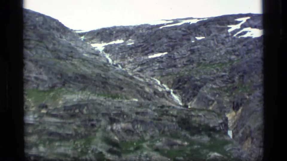 1977 Alaska aerial footage frame showing gray ridges, white snow streaks, green patches, and a narrow stream or waterfall.