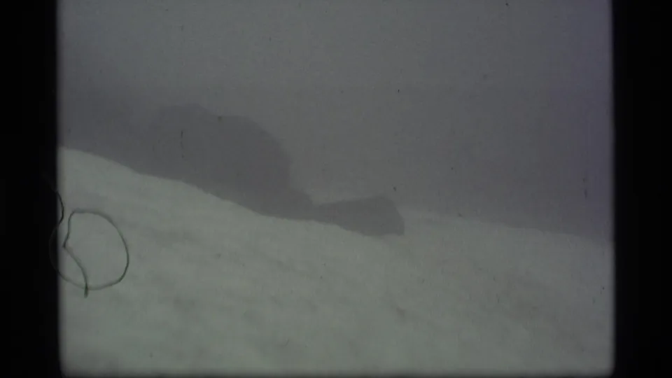 Faded 1977 Alaska home-movie frame with pale snowfield, dark mountain slope, cloudy haze, and visible 8mm film wear.