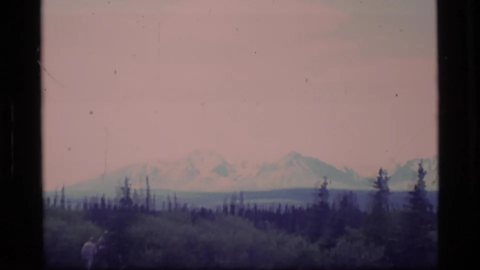 1977 Alaska 8mm frame showing snow-capped mountains behind evergreen forest, with a small distant person near the lower left edge.