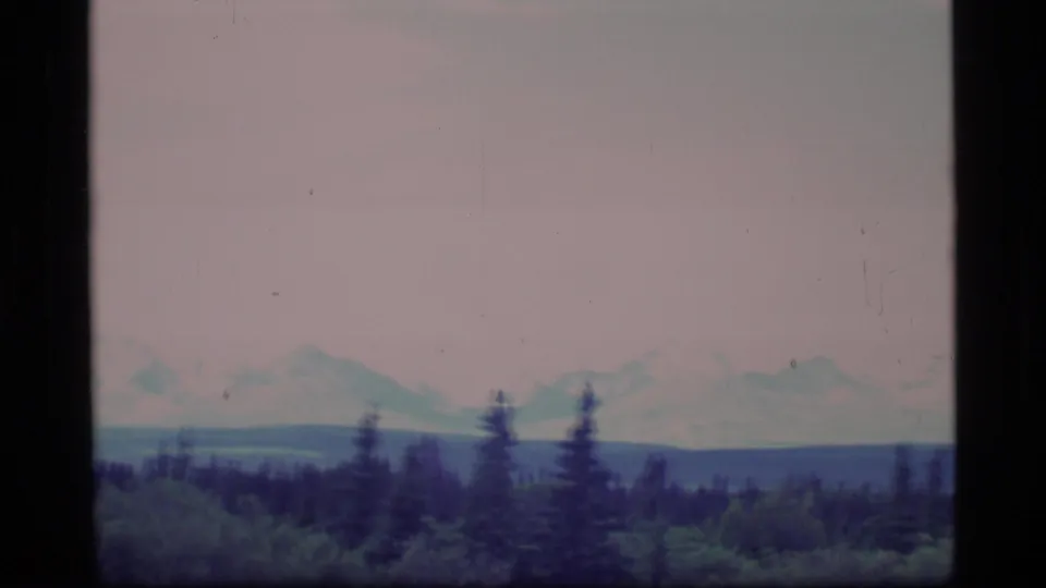 Faded 1977 Alaska home-movie frame showing a muted mountain range beyond evergreen forest and a pale sky.