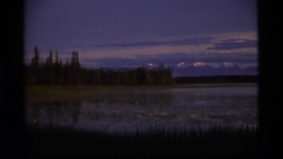 1977 Alaska 8mm dusk view across a calm lake toward a tree line, layered clouds, and distant mountain silhouettes.
