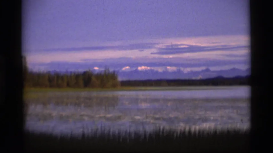 1977 Alaska 8mm frame of a still lake reflecting twilight color, with shoreline trees and low mountains in the distance.