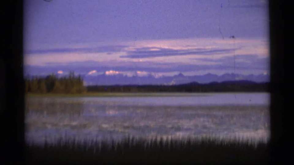 1977 Alaska 8mm wide dusk landscape with foreground grasses, a calm lake, distant shore trees, clouds, and mountain silhouettes.