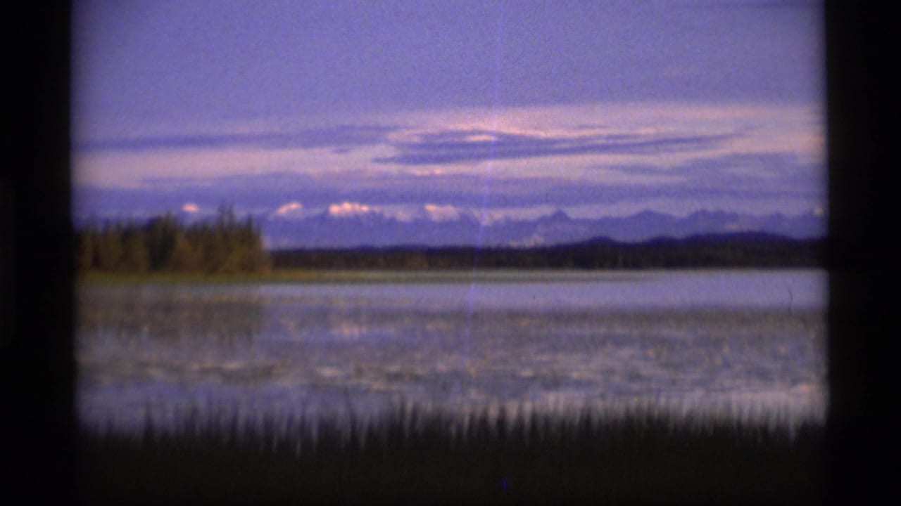 1977 Alaska 8mm frame showing a still lake at dusk with foreground reeds, a low treeline, distant mountains, and a purple-toned sky.