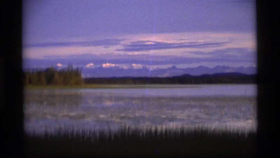 1977 Alaska 8mm frame showing a still lake at dusk with foreground reeds, a low treeline, distant mountains, and a purple-toned sky.