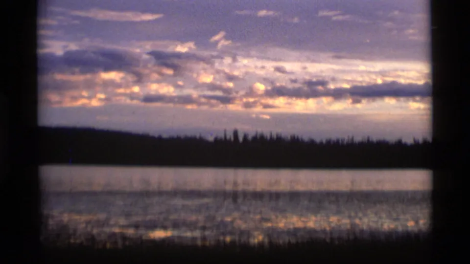1977 Alaska 8mm frame of a warm sunset over still water, with a dark treeline and glowing clouds reflected across the lake.