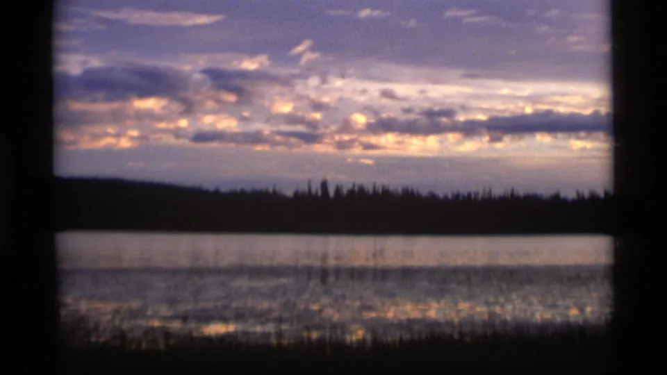 1977 Alaska 8mm frame holding on a lake at sunset, with glowing clouds above a silhouetted forest and reflected light near foreground reeds.