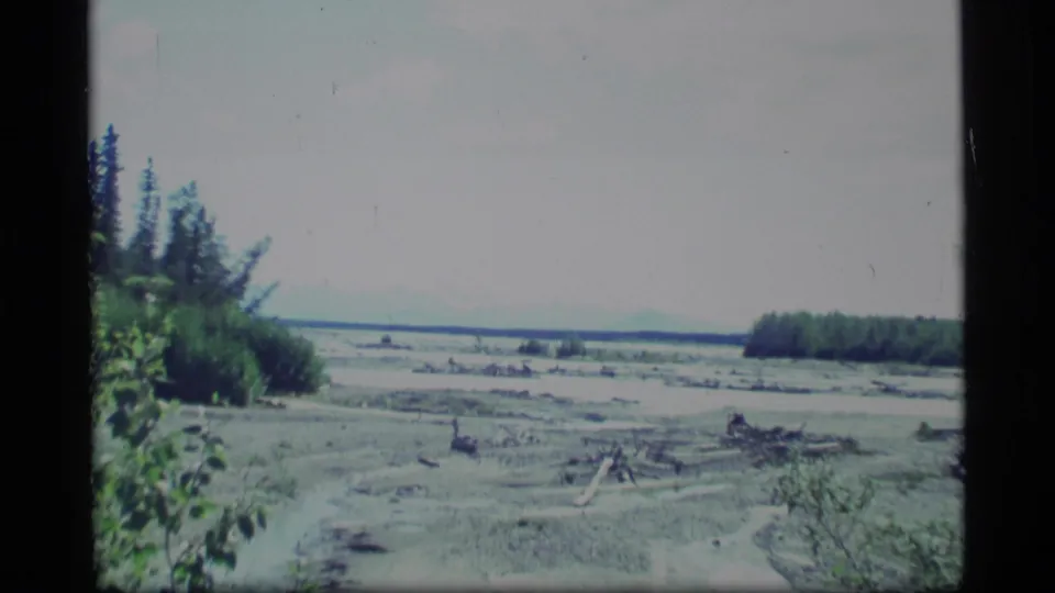 1977 Alaska 8mm frame showing a bright river delta or broad shoreline with driftwood, low vegetation, distant trees, and faint mountains.
