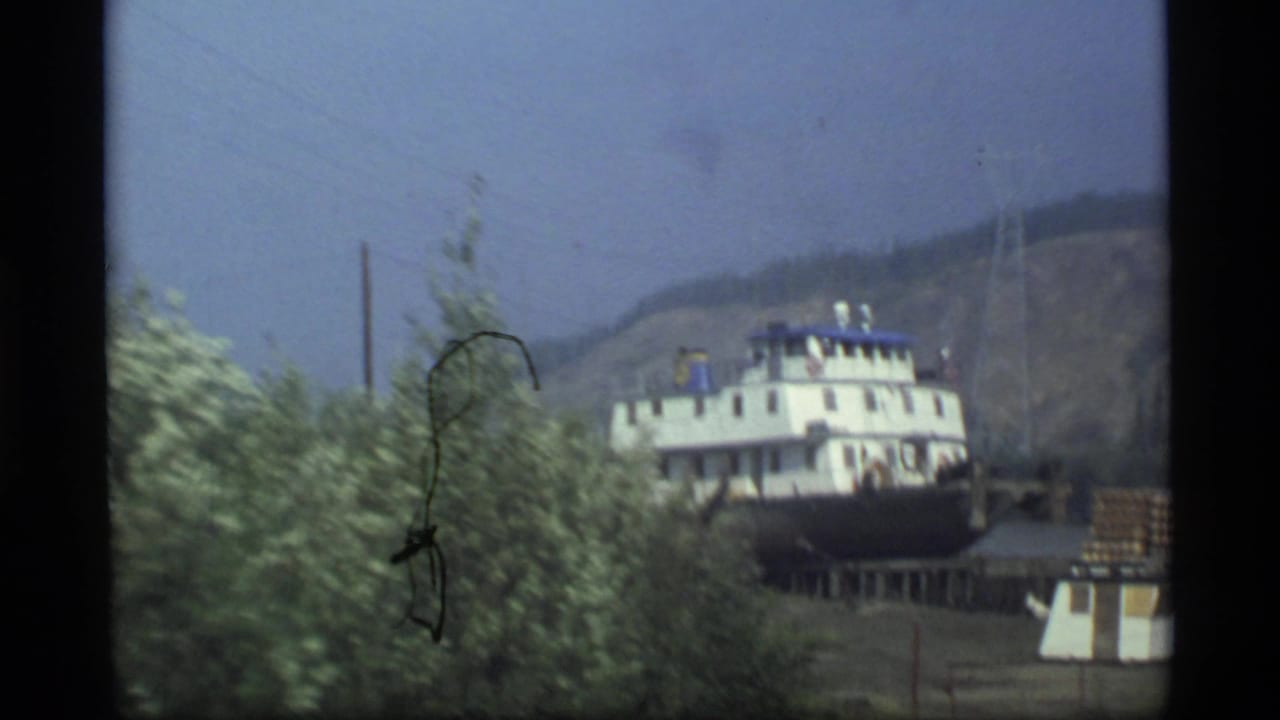 1977 Alaska 8mm frame showing a white and blue riverboat viewed through leafy trees with a hazy hillside behind it.