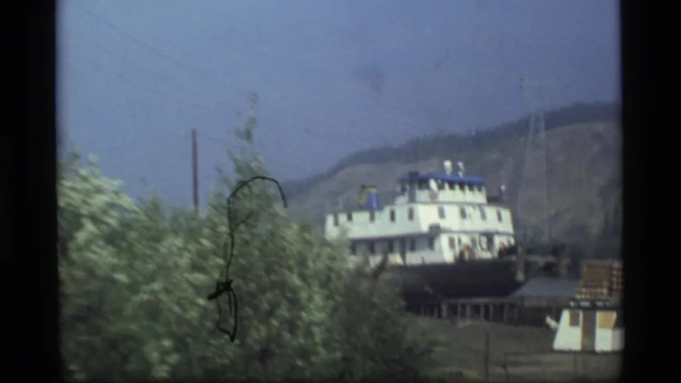 1977 Alaska 8mm frame showing a white and blue riverboat viewed through leafy trees with a hazy hillside behind it.