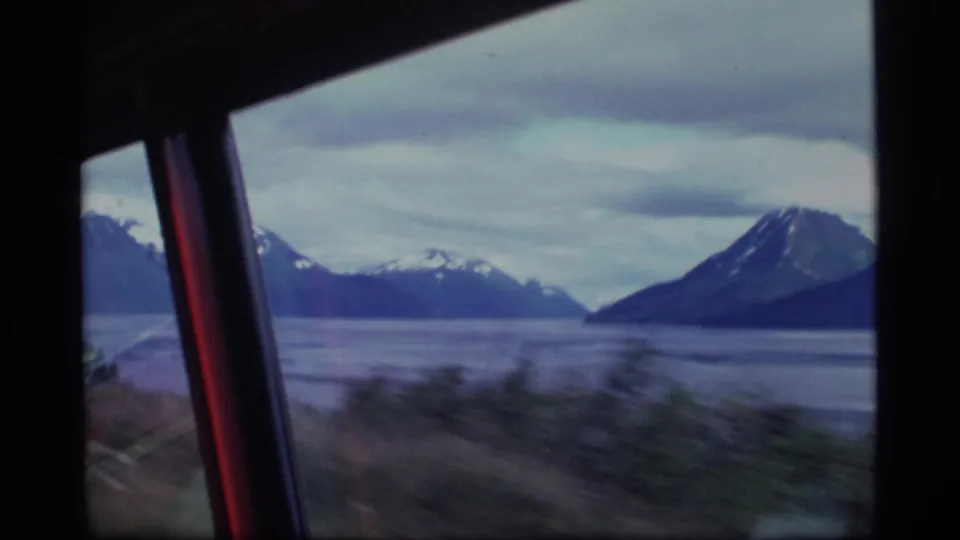 From inside a moving vehicle, 1977 Alaska 8mm footage frames a broad body of water and snowy mountains through the window.