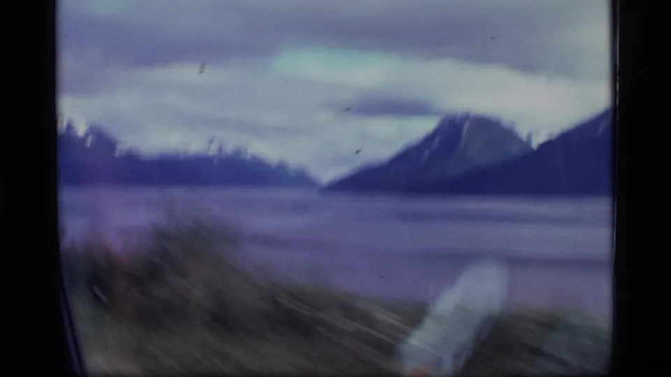 A moving-vehicle view in 1977 Alaska 8mm footage shows blurred foreground brush, calm water, and dark mountain forms.