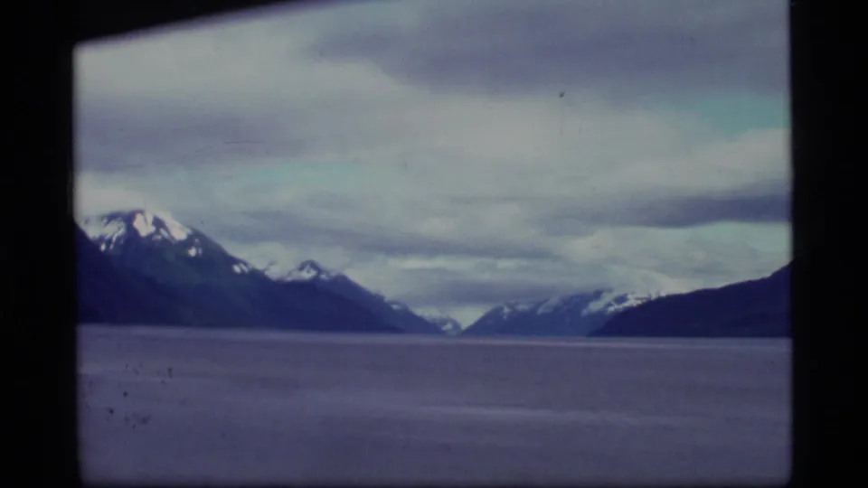 1977 Alaska 8mm footage shows snow-capped mountains beyond still water, with a dark shoreline edge and heavy overcast clouds.