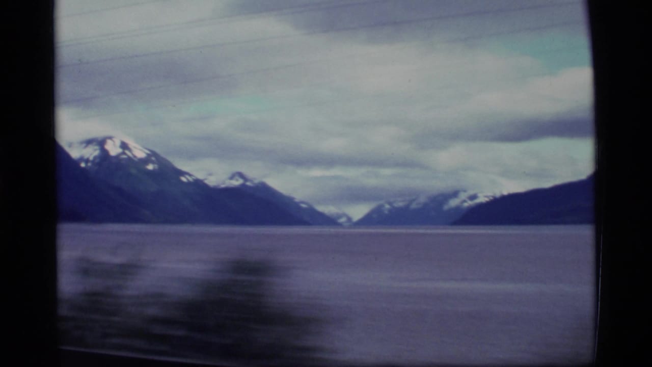 1977 Alaska 8mm travel view of calm water and distant snow-capped mountains framed by a dark window edge.